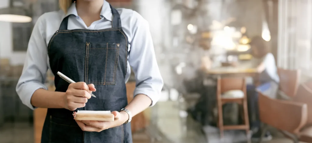 Restaurant server in an apron holding an order notepad during service in an Indian restaurant